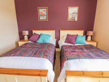 A bedroom with two single beds with colorful throws and pillows bedside tables with lamps and framed pictures on a purple wall at Cadwgan in Aberporth