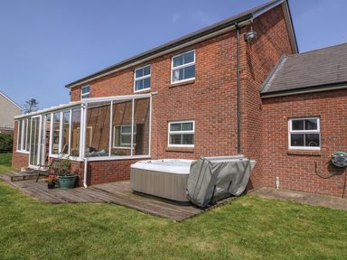 A backyard with a wooden deck a hot tub a red brick house with white windows and a glass sunroom at Cadwgan in Aberporth