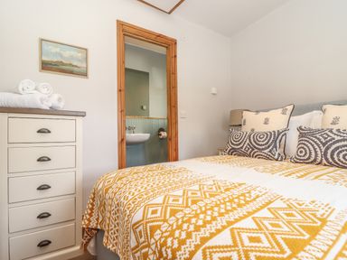 A bedroom with a bed with patterned blanket and pillows a chest of drawers with towels and a bathroom visible through the doorway at Swallows Nest in Morvah