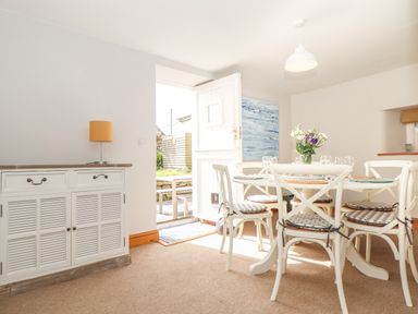 A dining room with a round table set with plates and glasses six chairs and a cabinet with a lamp near an open door at Swallows Nest in Morvah