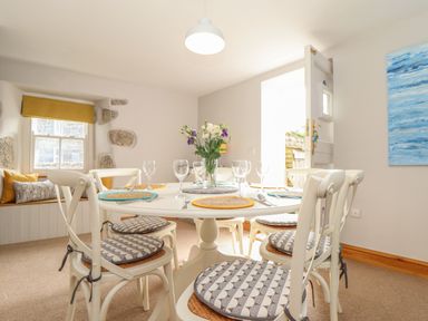 A dining room with a round table set with placemats and glasses chairs with cushions and a window seat with pillows at Swallows Nest in Morvah