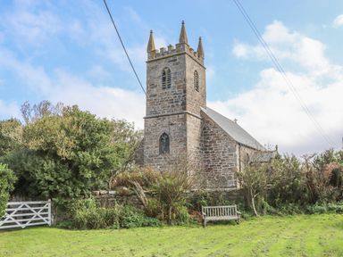 A stone church with a tower and battlements surrounded by bushes a wooden bench and a white gate at Swallows Nest in Morvah