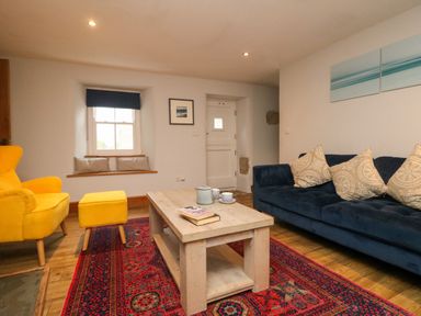A living room with a yellow armchair and footstool a blue sofa with cushions a wooden coffee table and a patterned rug at Swallows Nest in Morvah