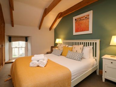 A bedroom with a bed with yellow throw and pillows beside a window and bedside tables with lamps at Swallows Nest in Morvah
