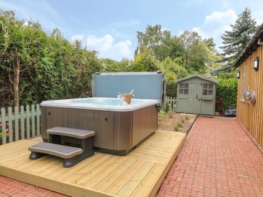 An outdoor area with a hot tub on a wooden deck a small green shed and a brick pathway at The Hen House in Stockwell Heath near Rugeley