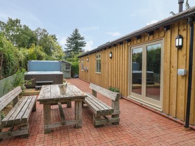 An outdoor patio area with wooden benches and table and a hot tub by a wooden building at The Hen House in Stockwell Heath near Rugeley