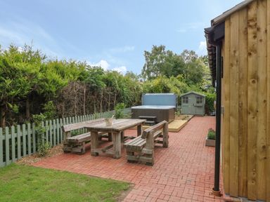 An outdoor patio area with wooden picnic table and benches a hot tub a small garden shed and greenery at The Hen House in Stockwell Heath near Rugeley