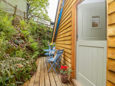 A narrow wooden deck with a half open door beside a small blue table and two blue chairs with cushions and plants nearby at Orchard Loft in Brownston near Modbury