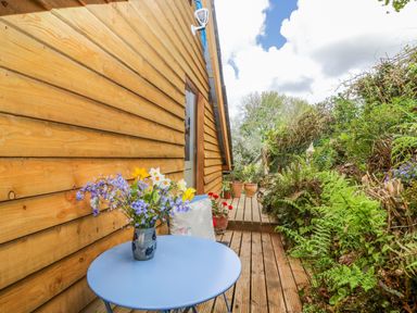 A small wooden deck with a blue round table holding a vase of flowers next to a wooden building and garden at Orchard Loft in Brownston near Modbury