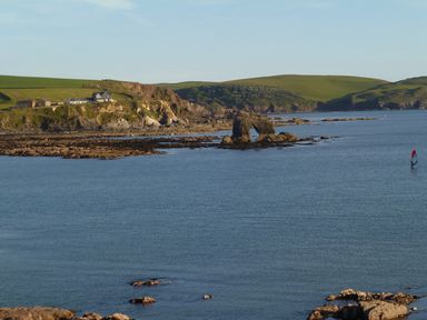 A coastal scene with rocky cliffs green hills houses on a hill and a person windsurfing in the water at Orchard Loft in Brownston near Modbury