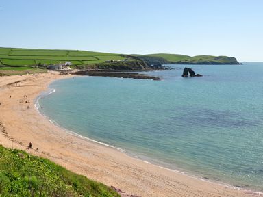 A sandy beach with people walking near the water and green fields with houses on cliffs overlooking the sea
