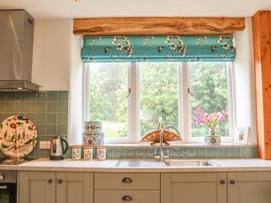 A kitchen countertop with a sink kettle and decorative containers by a window with a floral blind at The Old Laundry in North Tawton