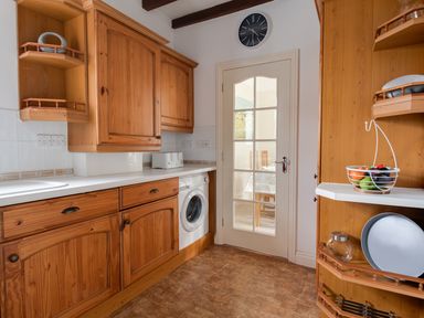 A kitchen with wooden cabinets white countertops a washing machine a toaster a clock on the wall and a fruit basket on a shelf at Nuke in Weymouth