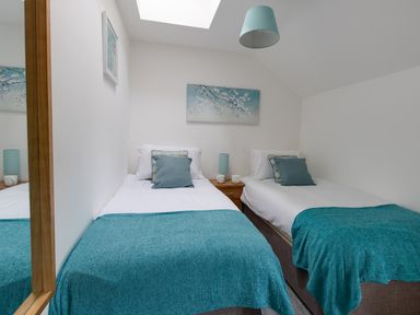 A bedroom with two single beds with blue throws and pillows a bedside table with lamps and mugs and wall art at Nuke in Weymouth