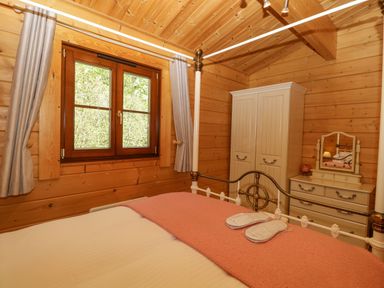 A bedroom with wooden walls a window a bed with a pink blanket and slippers a wardrobe and a dressing table with a mirror at Heron Lodge in Badwell Ash