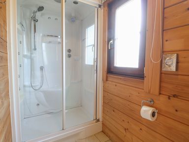 A corner shower enclosure with glass doors next to a wooden wall with a window and a toilet paper holder at Heron Lodge in Badwell Ash