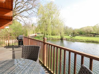 A wooden deck with a table, chairs, and a barbecue next to a pond with trees and grassy areas at Heron Lodge in Badwell Ash