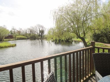A view of a pond with trees and a wooden railing with outdoor chairs at Heron Lodge in Badwell Ash