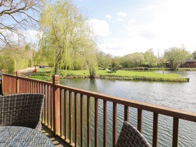A wooden deck with wicker chairs overlooking a pond and a grassy area with trees at Heron Lodge in Badwell Ash
