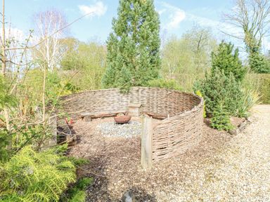A circular woven fence area with a fire pit in the center surrounded by trees and gravel at Heron Lodge in Badwell Ash