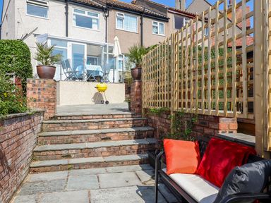 A backyard patio with stone steps a grill a table with chairs and cushions on a bench at Heddfan in Conwy