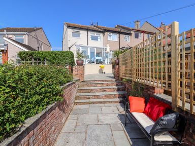A backyard patio area with stone steps leading to a raised patio with a table and chairs a bench with red and orange cushions and houses in the background at Heddfan in Conwy