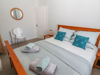 A bedroom with a wooden bed frame blue and grey bedding a white chair round wall mirror and wooden nightstand at Heddfan in Conwy