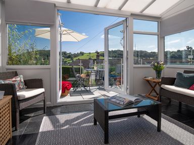 A sunroom with wicker furniture and a coffee table opening to a patio with outdoor chairs and umbrella at Heddfan in Conwy