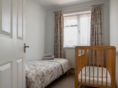 A small bedroom with a single bed covered with patterned bedding and a wooden baby crib near a window with patterned curtains at Heddfan in Conwy