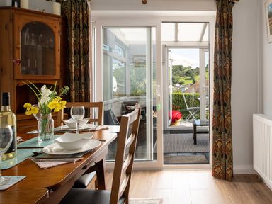 A dining area with a wooden table set with bowls glasses and a vase of flowers next to sliding glass doors leading to a patio with chairs and a table at Heddfan in Conwy