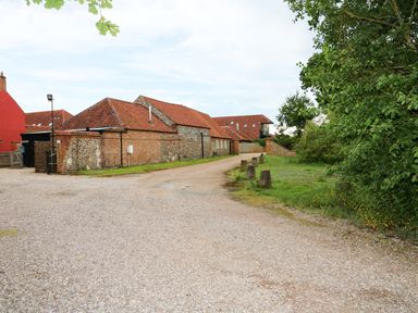 A gravel driveway next to brick and stone buildings with red roofs and greenery at Far Barn in Fakenham