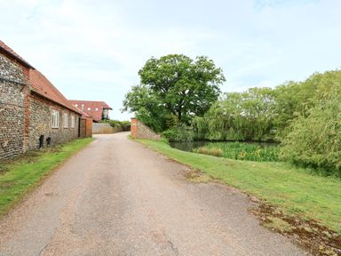 A gravel driveway between a stone house and a pond with trees at Far Barn in Fakenham