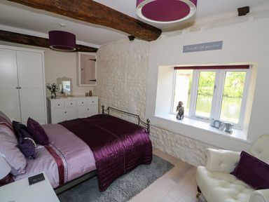 A bedroom with a bed purple bedspread white chest of drawers window with decorations and an armchair at Far Barn in Fakenham