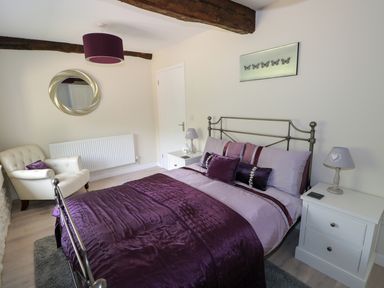 A bedroom with a metal bed frame purple bedding two white nightstands lamps a white armchair and a wall mirror at Far Barn in Fakenham