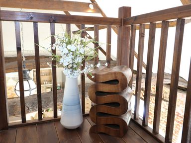 A wooden balcony railing with a white vase of flowers and a wooden decorative sculpture at Far Barn in Fakenham