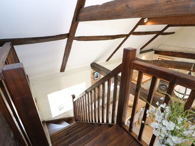 A wooden staircase with exposed ceiling beams and a dining area with flowers and appliances below at Far Barn in Fakenham