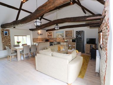 An open-plan living and kitchen area with wooden beams dining table and chairs beige sofas refrigerator and TV at Far Barn in Fakenham
