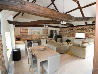 An open plan kitchen and living room with wooden beams a dining table with chairs beige sofas and a television at Far Barn in Fakenham
