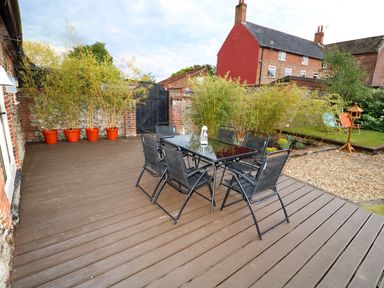 An outdoor patio with a glass table and six chairs potted plants along a wall and garden chairs in the background at Far Barn in Fakenham