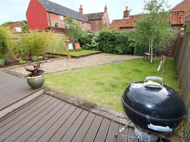 A backyard with wooden decking and a black barbecue grill a lawn with a tree and cricket equipment and a gravel path with deck chairs at Far Barn in Fakenham
