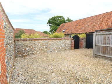 An outdoor gravel area enclosed by stone and brick walls with black wooden gates and tiled roof buildings at Far Barn in Fakenham
