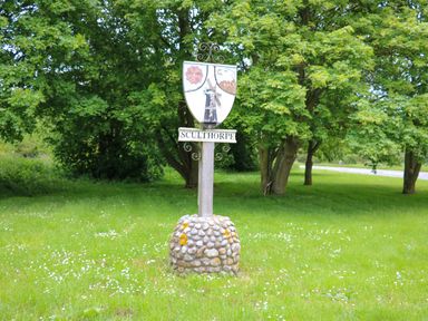 A village signpost with a shield and the name Sculthorpe on grass surrounded by trees