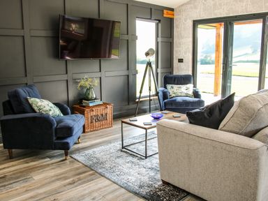 A living room with a television on the wall two blue armchairs a beige sofa a coffee table a wicker basket with books and flowers and sliding glass doors at Meadow View in Meifod Powys