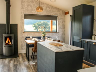 A kitchen with a marble countertop island a dining table with glasses and plates a wood burning stove and a window with a view at Meadow View in Meifod Powys