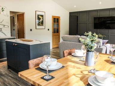 A dining table set with plates bowls and glasses next to a kitchen island and a living area with sofas and a wall mounted television at Meadow View in Meifod Powys