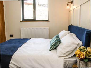 A bedroom with a bed with blue headboard and blanket with white pillows and a blue decorative pillow next to a window and a vase with yellow flowers at Meadow View in Meifod Powys