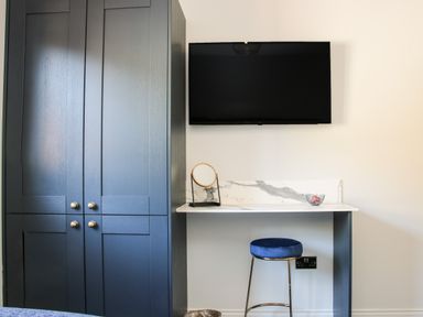 A room with a blue wardrobe a wall-mounted TV a white marble desk a blue stool and a small round mirror on the desk at Meadow View in Meifod Powys