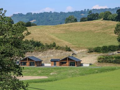 Two modern wooden houses with dark roofs on a grassy field with hills and trees in the background at Meadow View in Meifod Powys