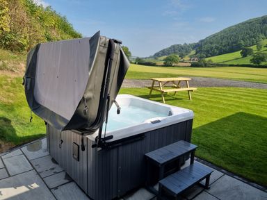 An outdoor hot tub with a partially open cover on a stone patio next to a wooden picnic table on grass at Meadow View in Meifod Powys