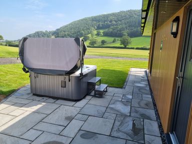 An outdoor patio with a hot tub covered by a partially open cover next to a wooden building and green hills in the background at Meadow View in Meifod Powys